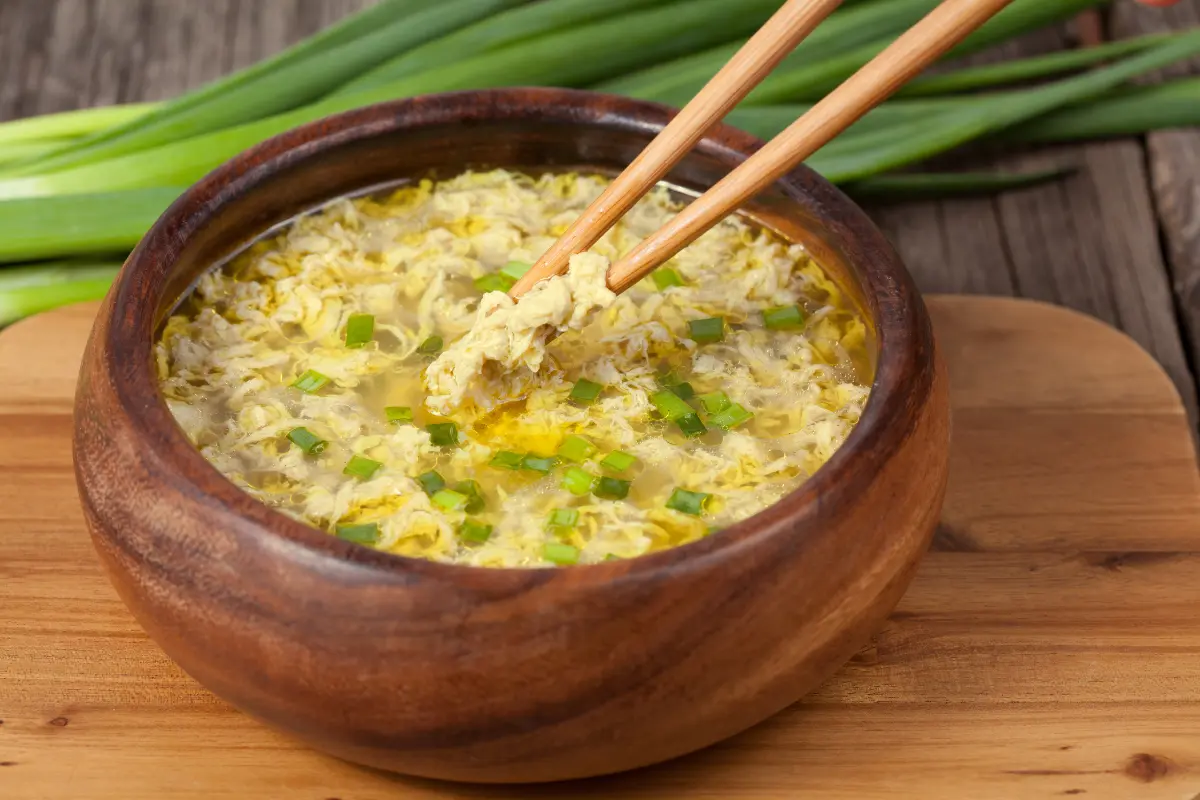 A steaming bowl of golden Chinese egg drop soup garnished with green onions and delicate egg ribbons, showcasing the simplicity and warmth of this traditional dish.