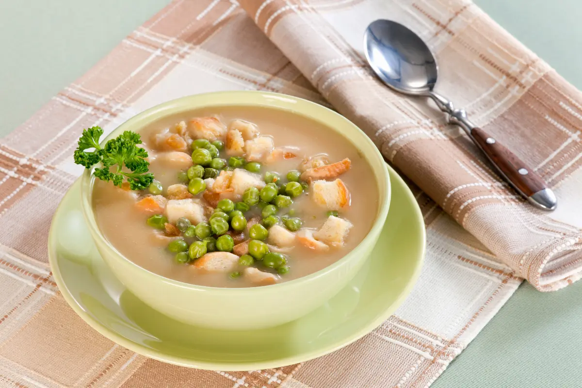 A cozy, steaming bowl of Anderson's pea soup placed on a rustic wooden table, surrounded by fresh peas, carrots, and vegan ingredients, highlighting its vegan-friendly recipe.