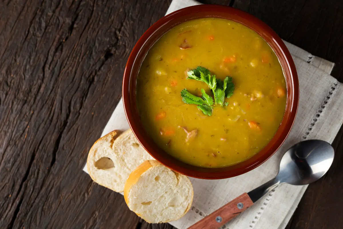 A cozy, steaming bowl of Anderson's pea soup placed on a rustic wooden table, surrounded by fresh peas, carrots, and vegan ingredients, highlighting its vegan-friendly recipe.