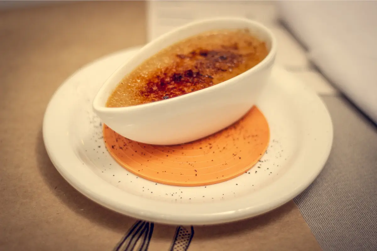 A close-up image showcasing a perfectly prepared crème brûlée with a golden caramelized top, a spoon gently breaking into the sugary crust to reveal the creamy custard beneath, set against a dark backdrop to highlight the dessert's texture and color contrast.